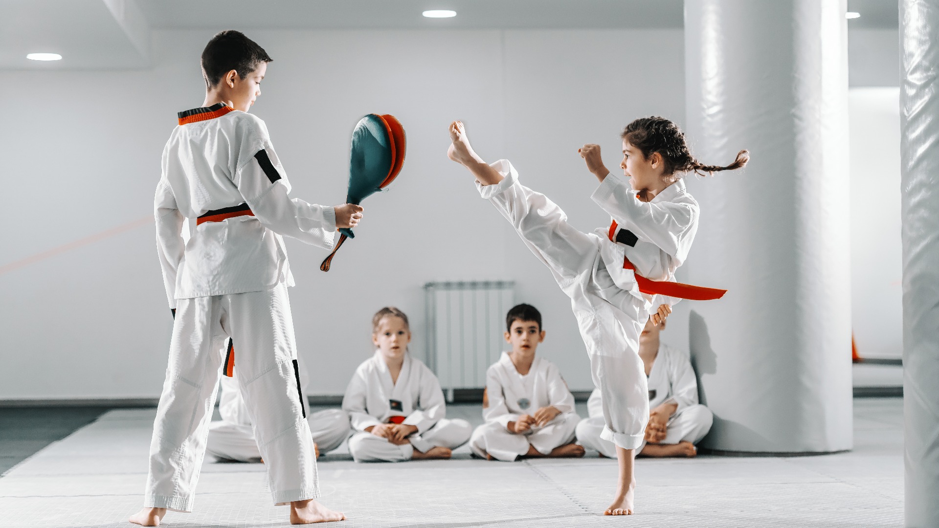 Caucasain boy and girl in doboks having taekwondo training at gym. Girl kicking while boy holding kick target. In background their friend sitting with legs crossed and watching them.