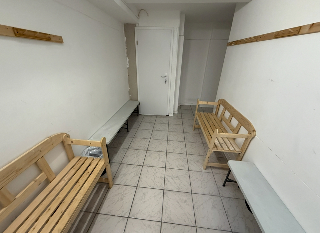 Umkleideraum mit Holzbänken, weiß gefliester Boden, weiße Wände, hölzerne Kleiderhaken.