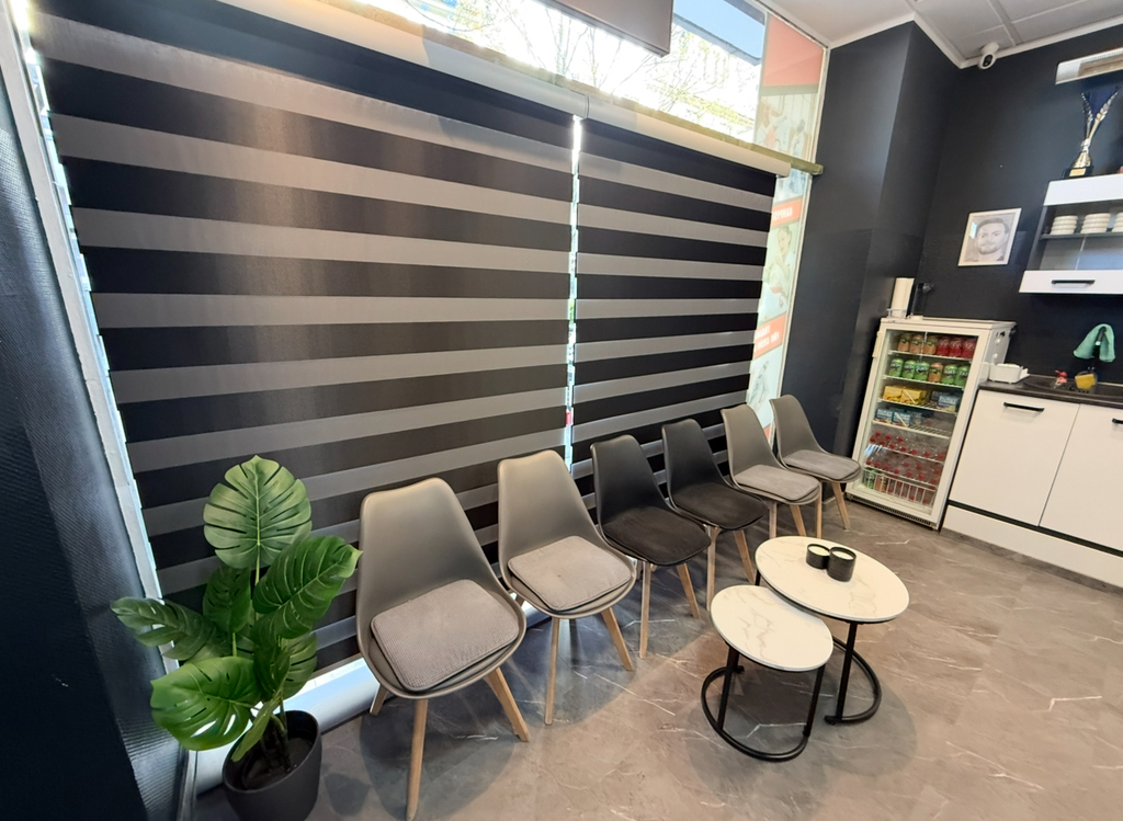 Warteraum mit grauen Stühlen, Zebra-Jalousien, Pflanze und Tisch auf grauen Fliesen.