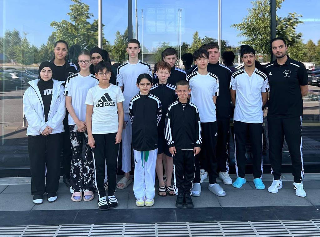 Eine Gruppe Jugendlicher und ein Erwachsener in sportlicher Kleidung vor einem Gebäude mit Glastür posieren lächelnd für ein Foto.